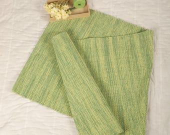 Può includere: Un tappeto tessuto a strisce verdi e gialle con un bordo ripiegato. Il tappeto è su una superficie bianca con una piccola scatola di legno che contiene un vaso verde e fiori bianchi.