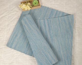 Puede incluir: Una alfombra redonda de color azul claro con un sutil patrón de rayas blancas. La alfombra está enrollada y parcialmente desenrollada, revelando la textura de la tela.
