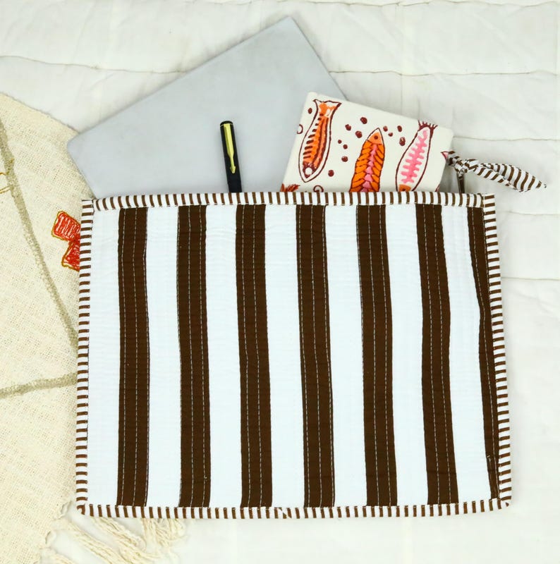 May include: A rectangular quilted pouch featuring vertical brown and white stripes with a matching striped border. Inside the pouch are a grey laptop, a black pen, and a white wallet decorated with a colourful orange and pink fish pattern.