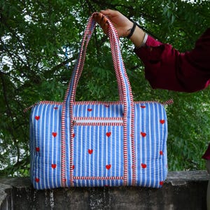 May include: A quilted duffel bag featuring a light blue and white vertical striped pattern with small red heart icons scattered across the surface. The bag includes two long shoulder straps and a front zippered pocket with red and white trim.