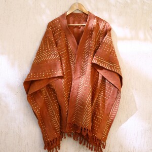 Puede incluir: Un kimono de color óxido con un patrón geométrico en un tono naranja más claro. El kimono tiene cuello en V y mangas largas con flecos en la parte inferior.