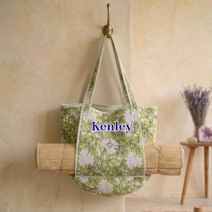 May include: A quilted tote bag featuring a green and white floral pattern with the name Kenley printed in blue on the front. The bag has two striped handles and holds a rolled tan textured mat horizontally across its center.