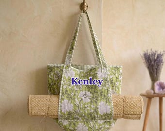 May include: A quilted tote bag featuring a green and white floral pattern with the name Kenley printed in blue on the front. The bag has two striped handles and holds a rolled tan textured mat horizontally across its center.