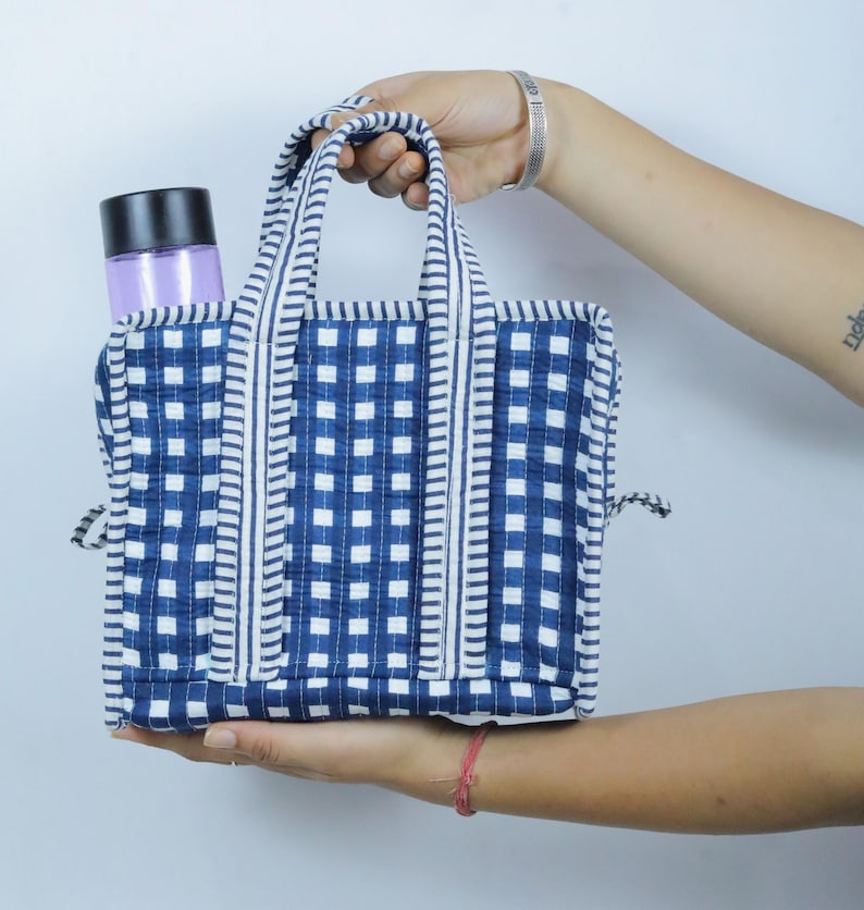 以下が含まれることがあります： 青と白のチェック柄の小さなトートバッグ。持ち手とトリムも同じ柄です。バッグは長方形で、黒い蓋が付いた紫色のボトルが入っています。バッグは人が持っています。