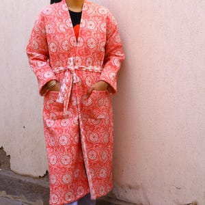Puede incluir: Una bata acolchada de color coral con un estampado floral blanco. La bata tiene mangas largas, un cinturón a juego y bolsillos. La bata llega a la rodilla y parece estar hecha de una tela suave y ligera.