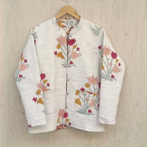 Könnte beinhalten: Eine weiße gesteppte Jacke mit einem Blumendruck mit rosa, roten und gelben Blumen. Die Jacke hat einen Stehkragen und einen Knopfverschluss.