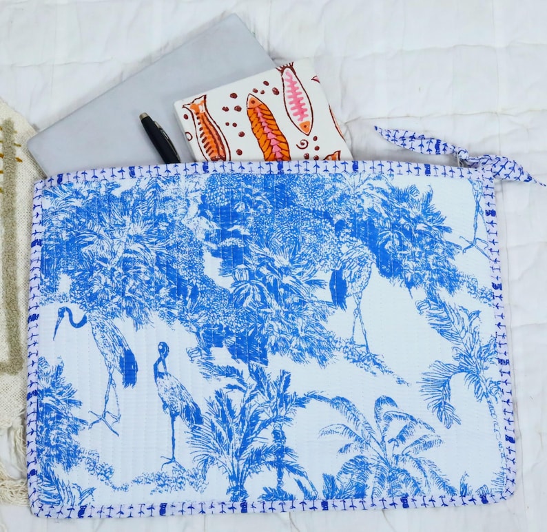 May include: A rectangular quilted laptop sleeve featuring a blue and white toile pattern of cranes and palm trees. The sleeve has a zip closure and contains a silver laptop, a black pen, and a white notebook with orange fish illustrations.