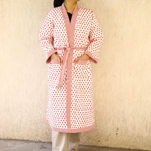 Könnte beinhalten: Ein weißer Bademantel mit rotem Polka-Dot-Muster. Der Bademantel hat eine gestreifte Bordüre und einen passenden Gürtel. Er hat Taschen und lange Ärmel. Der Bademantel ist knielang und aus einem weichen, bequemen Material.