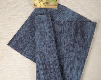 Puede incluir: Una alfombra tejida azul y gris con una superficie texturizada. La alfombra está doblada por la mitad y está sobre una superficie blanca.