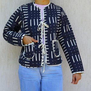May include: A navy blue jacket with a white vertical stripe pattern and two front pockets. The jacket has a round collar and a tie closure. The jacket is worn over a pink shirt and blue jeans.