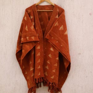 Könnte beinhalten: Ein rostfarbener Poncho mit einem geometrischen Muster aus Dreiecken. Der Poncho hat einen Fransenbesatz und ist aus einem weichen, gewebten Stoff gefertigt.