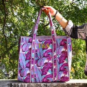 May include: A quilted tote bag with a vibrant tropical leaf pattern in shades of purple, blue, and white. The bag features long handles and a rectangular shape. The initials "T&S" are embroidered in gold on the front.