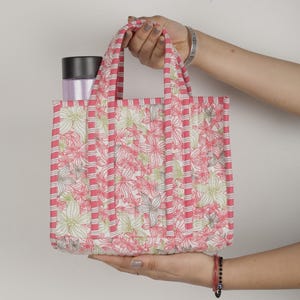 May include: A small, quilted tote bag with a floral pattern in pink, white, and green. The bag has pink striped accents and handles. A black and silver container is inside the bag.
