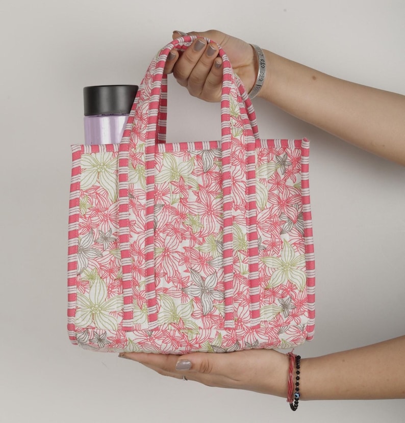 May include: A small, quilted tote bag with a floral pattern in pink, white, and green. The bag has pink striped accents and handles. A black and silver container is inside the bag.
