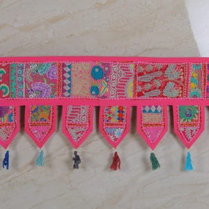 Op de afbeelding: Een roze stoffen deurhanger met kleurrijke patchwork-vierkanten en kwastjes. De hanger heeft een gegolfde rand en is versierd met borduurwerk.