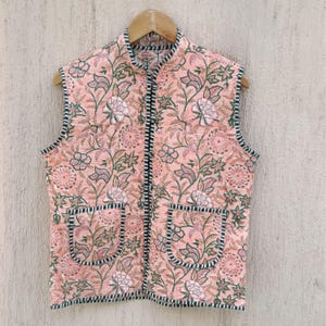 Könnte beinhalten: Eine ärmellose, pfirsichfarbene gesteppte Weste mit Blumendruck und schwarzem Besatz. Die Weste hat einen Reißverschluss und zwei Taschen.
