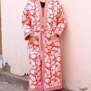 Op de afbeelding: Een lange, rood-witte bloemenpatroon badjas met bijpassende riem. De badjas heeft zakken en gestreepte randen langs de randen en mouwen. Het bloemenpatroon is een herhalend ontwerp van witte vormen op een rode achtergrond. De badjas wordt over een zwart shirt gedragen.