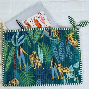 Può includere: Un sottopiatto trapuntato con una scena della giungla con donne, leopardi e un fogliame lussureggiante in tonalità di verde e blu. Il sottopiatto ha un bordo a righe verdi e bianche. Sono visibili un taccuino, una penna e una pochette fantasia.