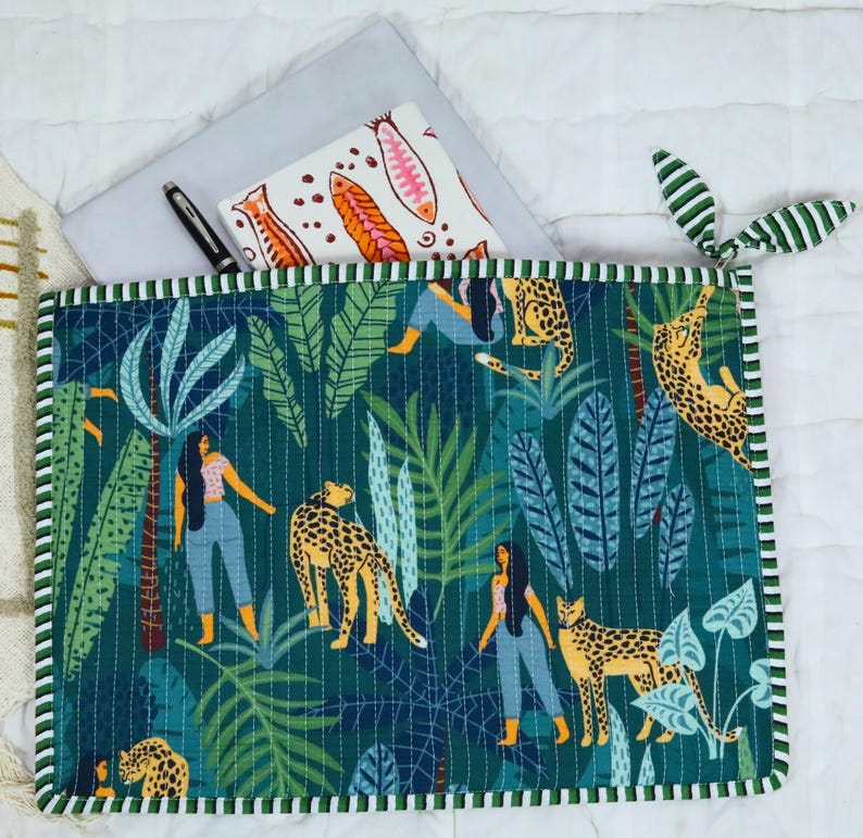 May include: A rectangular quilted laptop sleeve featuring a dark green jungle print with illustrations of leopards and people in blue jeans. The sleeve has a striped green and white border, a zip closure with fabric tabs, and contains a notebook.