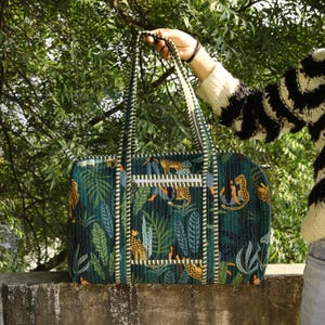 May include: A rectangular quilted duffel bag featuring a dark green jungle print with leopards and tropical foliage. The bag has two long shoulder straps with a white and green striped pattern, a front zippered pocket, and a top zipper closure.