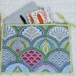 May include: A rectangular quilted fabric sleeve featuring a repeating fan-shaped pattern in shades of blue, green, pink, and white. The sleeve has a light green and white striped border and contains a grey laptop, a black pen, and a white notebook.
