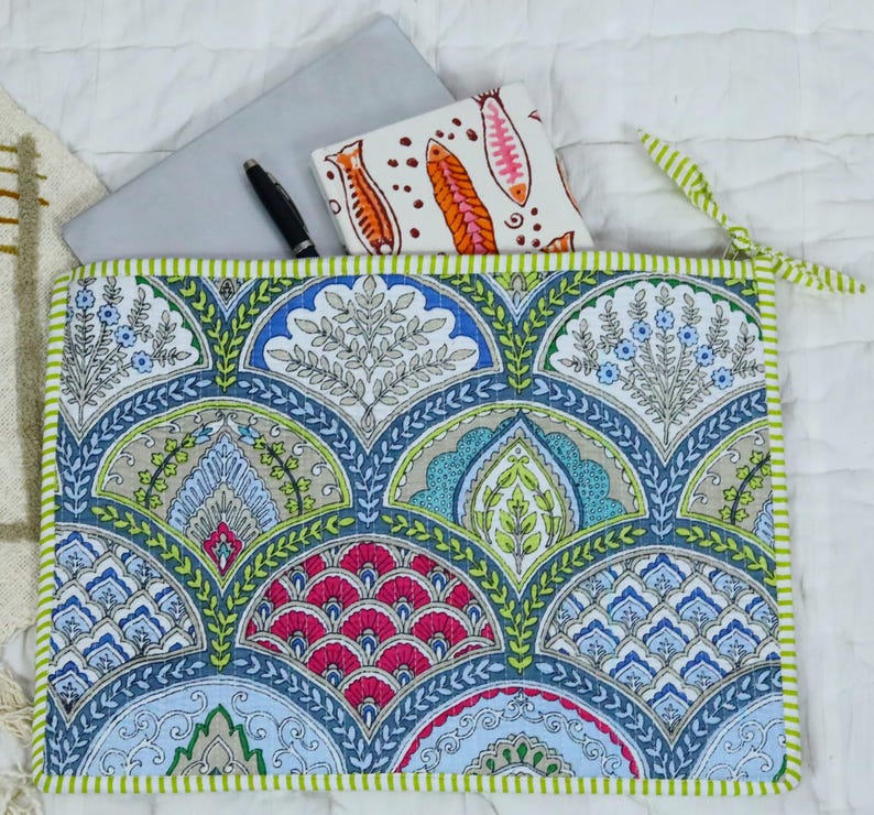 May include: A rectangular quilted fabric sleeve featuring a repeating fan-shaped pattern in shades of blue, green, pink, and white. The sleeve has a light green and white striped border and contains a grey laptop, a black pen, and a white notebook.
