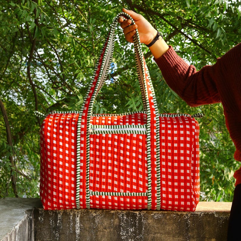May include: A rectangular quilted duffel bag featuring a vibrant red and white gingham pattern. The bag includes two long shoulder straps, a horizontal zippered front pocket, and green and white striped fabric trim along the edges and handles.