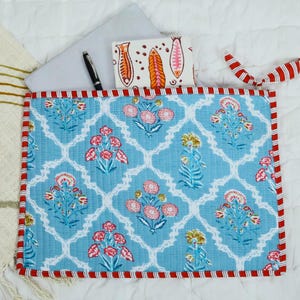 May include: A quilted fabric laptop sleeve featuring a light blue background with a white lattice pattern and floral motifs in pink, red, and yellow. The sleeve has red and white striped trim, a black pen, and a small notebook with fish illustrations.
