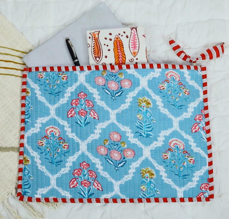 May include: A quilted fabric laptop sleeve featuring a light blue background with a white lattice pattern and floral motifs in pink, red, and yellow. The sleeve has red and white striped trim, a black pen, and a small notebook with fish illustrations.