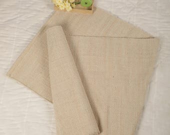 Può includere: Un tappeto beige tessuto con una superficie testurizzata. Il tappeto è piegato a metà e si trova su una superficie bianca.