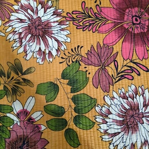 May include: A quilted fabric featuring a mustard yellow background with a repeating floral pattern. The design includes large white and magenta chrysanthemums, smaller pink blossoms, and green leafy branches with visible vertical stitching lines.