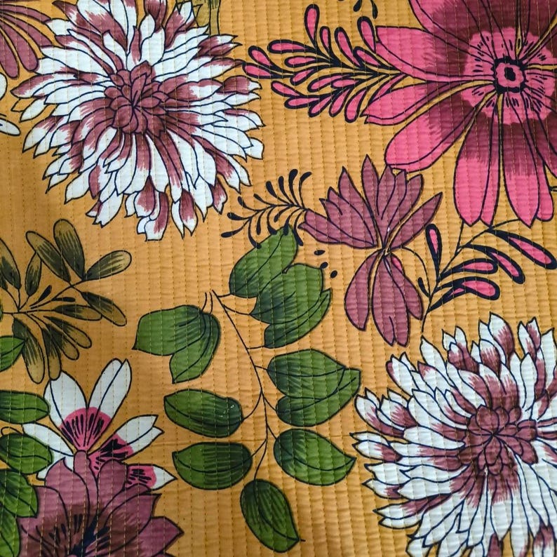 May include: A quilted fabric featuring a mustard yellow background with a repeating floral pattern. The design includes large white and magenta chrysanthemums, smaller pink blossoms, and green leafy branches with visible vertical stitching lines.