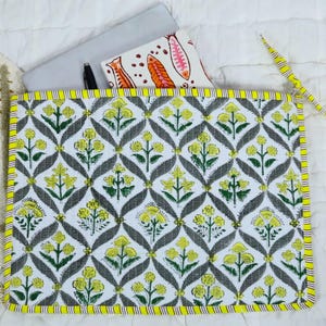 May include: A rectangular quilted laptop sleeve featuring a repeating pattern of yellow flowers and green leaves set against a white background with grey diamond-shaped outlines. The sleeve has a bright yellow and white striped fabric border.