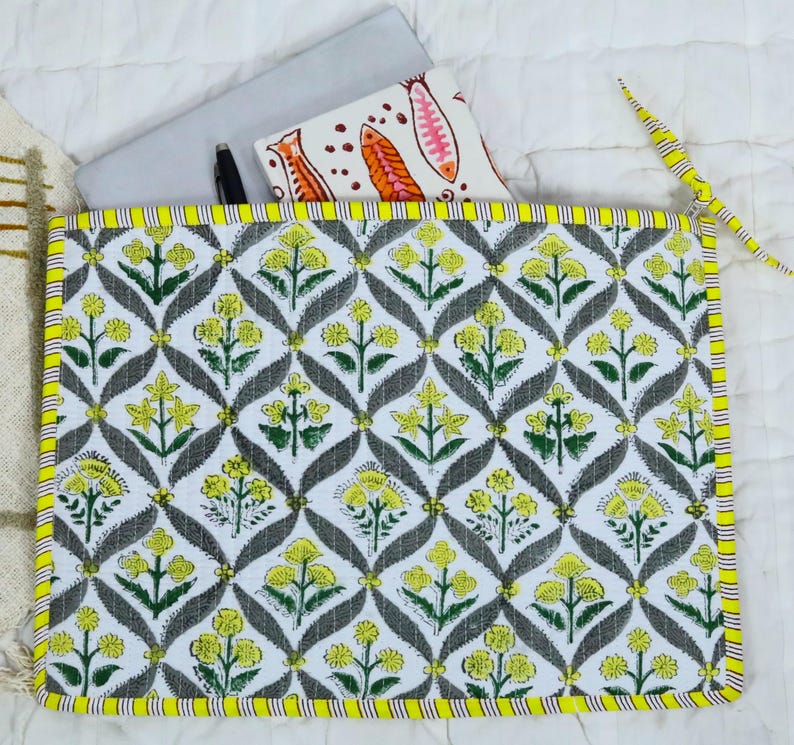 May include: A rectangular quilted laptop sleeve featuring a repeating pattern of yellow flowers and green leaves set against a white background with grey diamond-shaped outlines. The sleeve has a bright yellow and white striped fabric border.