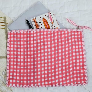 May include: A rectangular quilted fabric pouch featuring a red and white gingham pattern with a matching fabric zip pull. Inside the pouch, a silver laptop and a white notebook with an orange and pink fish print are partially visible.