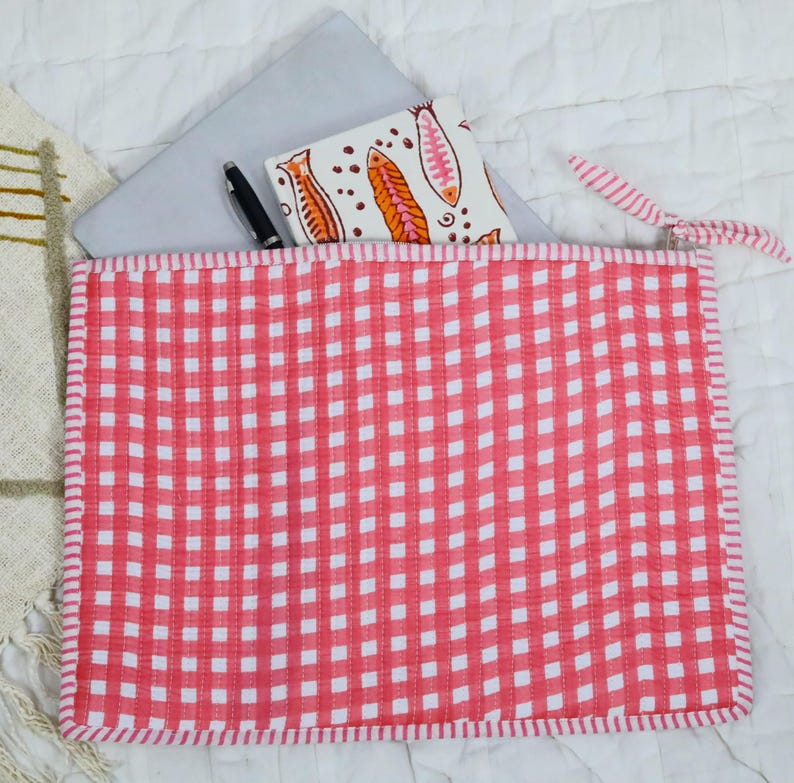 May include: A rectangular quilted fabric pouch featuring a red and white gingham pattern with a matching fabric zip pull. Inside the pouch, a silver laptop and a white notebook with an orange and pink fish print are partially visible.