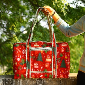 May include: A quilted duffel bag featuring a vibrant red background with a repeating pattern of green Christmas trees, wrapped gift boxes, and candy canes. The bag includes two long shoulder straps and a front zippered pocket with green and white stripes.