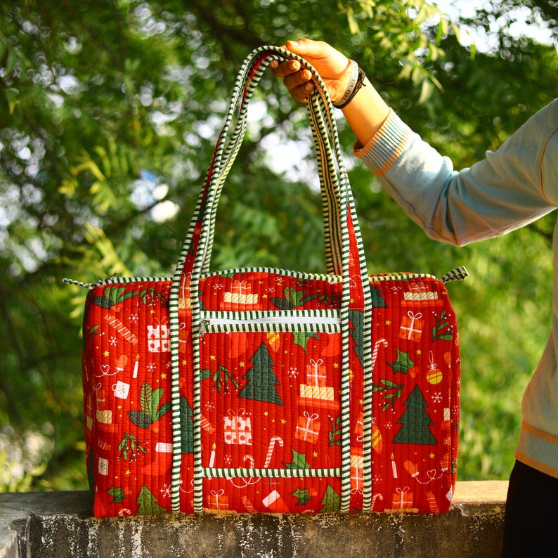 May include: A quilted duffel bag featuring a vibrant red background with a repeating pattern of green Christmas trees, wrapped gift boxes, and candy canes. The bag includes two long shoulder straps and a front zippered pocket with green and white stripes.