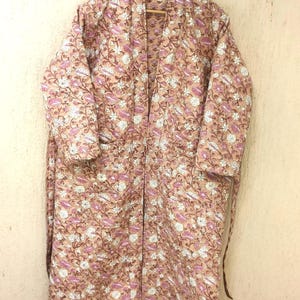 May include: A long-sleeved, quilted robe featuring a light brown base color with an all-over floral pattern in shades of white and pink. The garment has a shawl collar, an open front design, and a matching fabric tie belt at the waist.