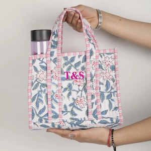 May include: A white tote bag with a floral pattern in pink and blue, with the letters "T&S" in pink. The bag has pink handles and trim, and contains a purple water bottle. The bag is held by a person.
