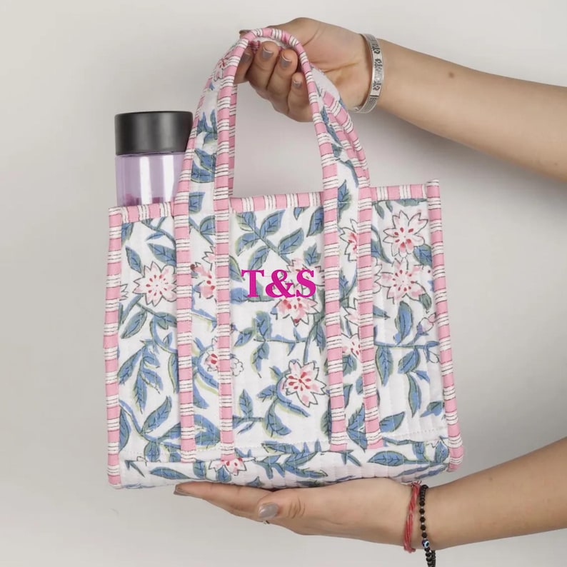 May include: A white tote bag with a floral pattern in pink and blue, with the letters "T&S" in pink. The bag has pink handles and trim, and contains a purple water bottle. The bag is held by a person.