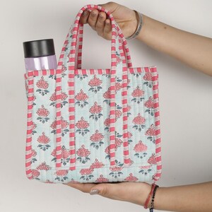 May include: A quilted tote bag featuring a floral design in pink and light blue. The bag has pink striped handles and trim. A water bottle is in the side pocket. The bag is held by a person.