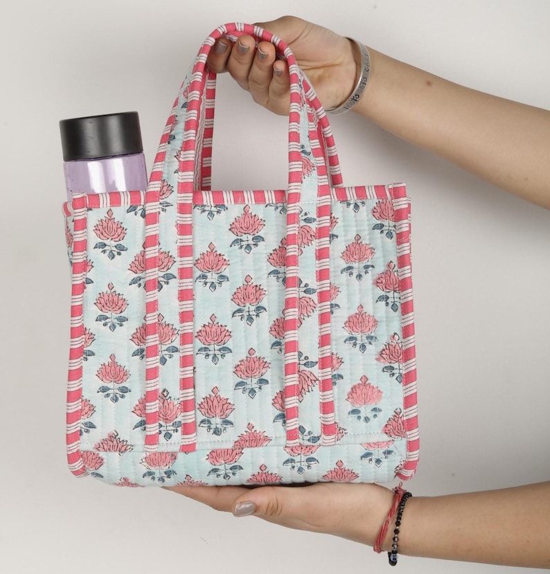 May include: A quilted tote bag featuring a floral design in pink and light blue. The bag has pink striped handles and trim. A water bottle is in the side pocket. The bag is held by a person.