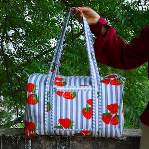 May include: A quilted duffel bag featuring a pattern of red strawberries on a white and light blue vertical striped background. The bag includes two long shoulder straps, a zippered top closure, and a front exterior pocket with a zipper.