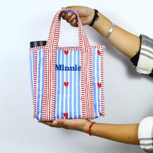 May include: A small tote bag with red and white stripes and blue vertical stripes. The bag has red heart accents and the name "Minnie" embroidered in blue. The handles are red and white striped.