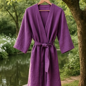 May include: A purple cotton robe with a tie belt hanging on a wooden hanger. The robe is hanging from a tree branch over a pond.