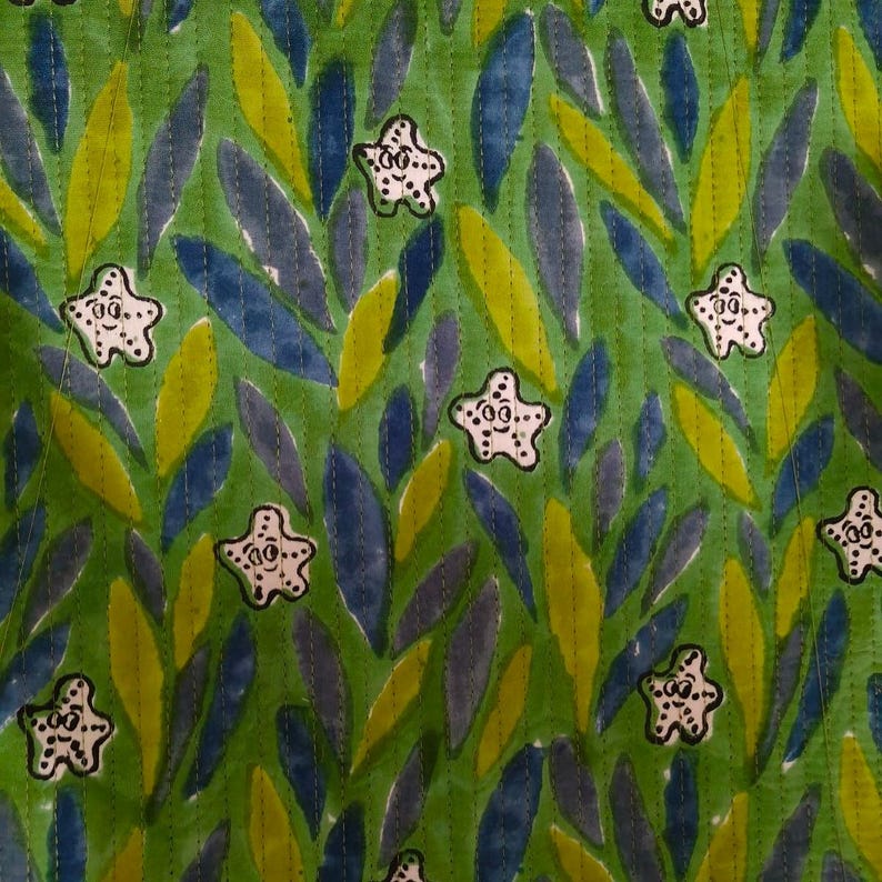 May include: A quilted fabric featuring a repeating pattern of white starfish with black outlines and facial features, set against a background of overlapping green and blue leaf-like shapes, with vertical stitching lines running across the textile.
