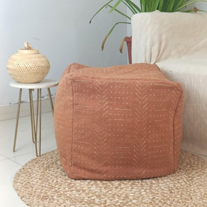 May include: A square, terracotta-colored ottoman with a geometric pattern in a lighter shade of brown. The ottoman is sitting on a woven rug.