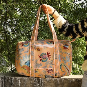 May include: A quilted duffel bag in a soft peach hue featuring a vibrant floral and paisley print in shades of blue, red, and green. The bag includes two long shoulder straps, a rectangular front pocket with a white zipper, and striped trim.