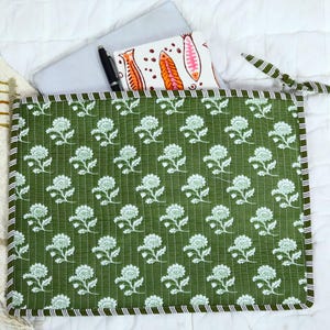 May include: A rectangular quilted laptop sleeve in olive green featuring a repeating white floral pattern. The sleeve has a striped border and a zip closure with a fabric tie. Inside, a silver laptop and a white notebook with fish illustrations.